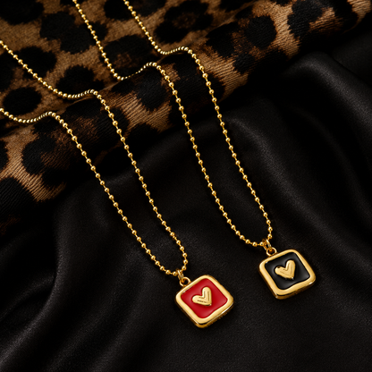 Two gold ball-chain necklaces with square pendant charms, one featuring a red background with a gold heart and the other with a black background and a gold heart, displayed on a black satin fabric with a leopard print textile.