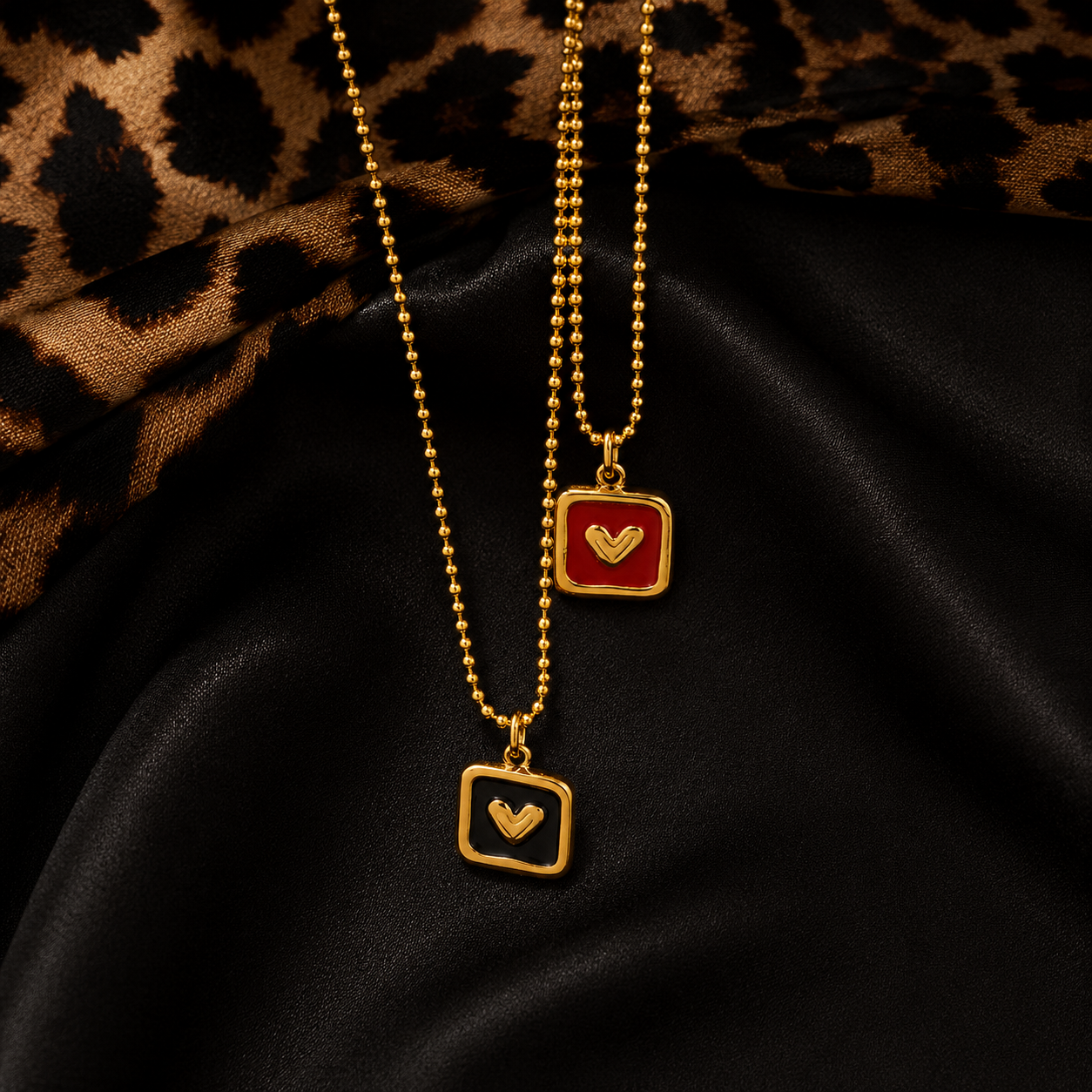 Two gold ball-chain necklaces with square pendant frames, one featuring a red enamel background and the other black, both with a small heart design in the center, displayed on a black satin fabric with a leopard print cloth in the background.
