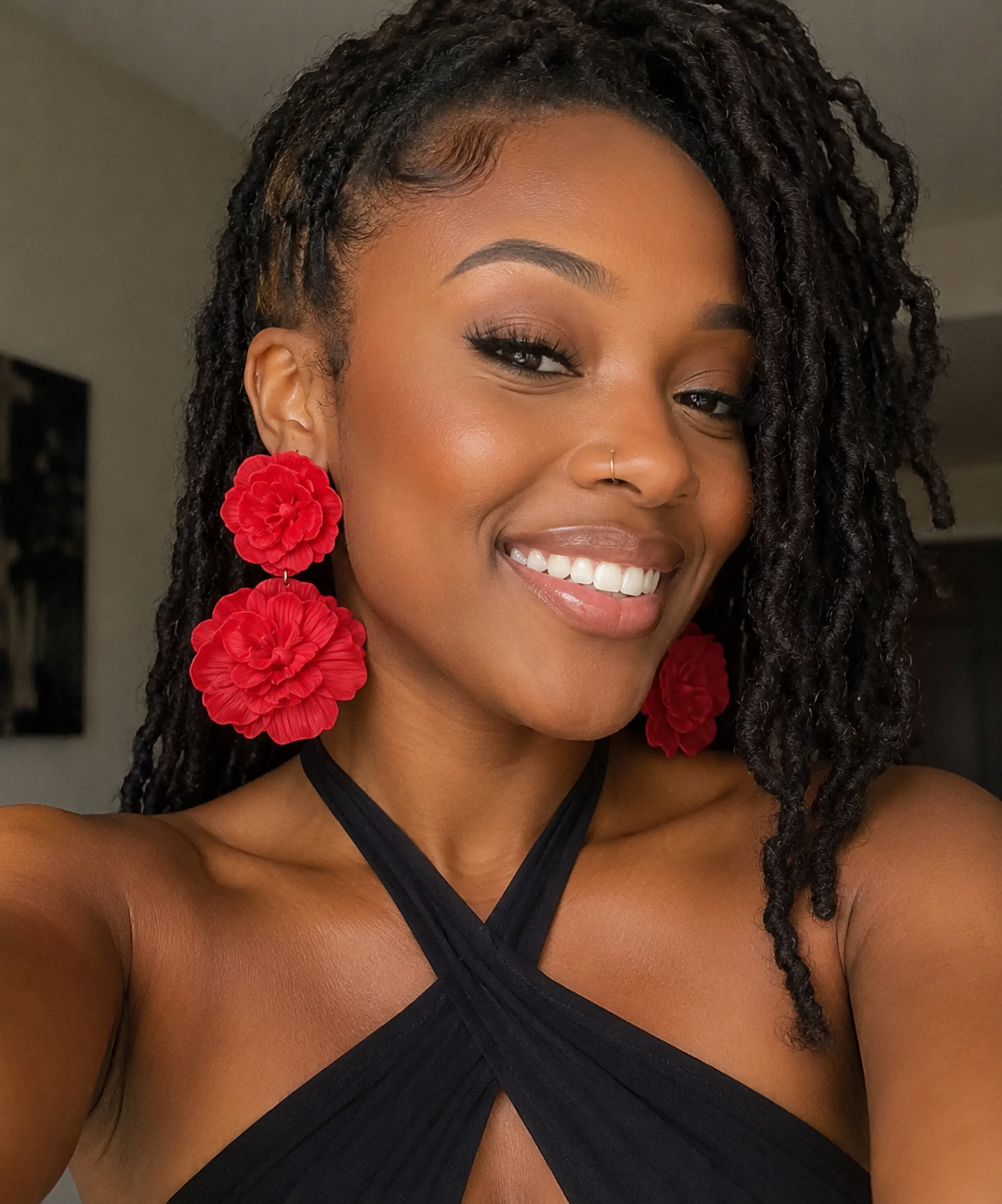 A smiling woman with dark dreadlocks wears large, vibrant red flower-shaped earrings and a black halter top in an indoor setting.